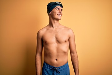 Poster - Young handsome man shirtless wearing swimsuit and swim cap over isolated yellow background looking away to side with smile on face, natural expression. Laughing confident.