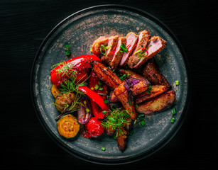 Wall Mural - fried meat with potatoes, pepper, tomatoes, herbs and spices in plate on black wooden table background