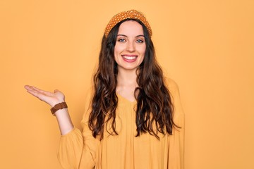 Wall Mural - Young beautiful woman wearing casual t-shirt and diadem standing over yellow background smiling cheerful presenting and pointing with palm of hand looking at the camera.