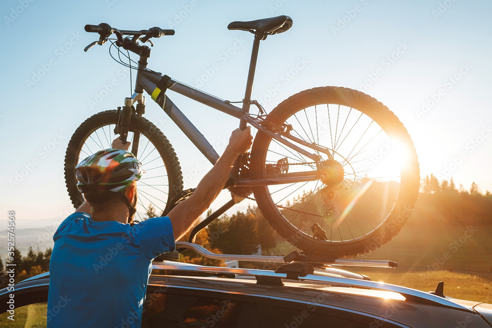 Young Man dresses modern cycling clothes and protective helmet installing his mountain bike on the car roof with sunset backlight. Active sporty people concept image. - obrazy, fototapety, plakaty 