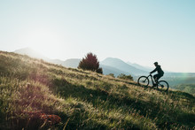Athletic Man Pedalling An MTB E-bike Up A Steep Grassy Hill. Beautiful View Of The Mountains At Sunrise/sunset With Sun Flare. Alone In Nature, Thinking About Life.