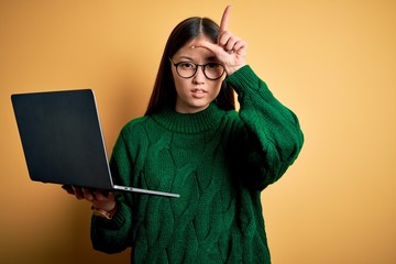 Poster - Young asian business woman wearing glasses and working using computer laptop making fun of people with fingers on forehead doing loser gesture mocking and insulting.