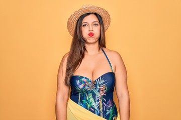 Sticker - Young hispanic woman wearing summer hat and swimsuit over yellow background puffing cheeks with funny face. Mouth inflated with air, crazy expression.