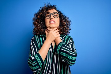 Poster - Young beautiful curly arab woman wearing striped shirt and glasses over blue background shouting and suffocate because painful strangle. Health problem. Asphyxiate and suicide concept.