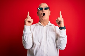 Wall Mural - Middle age hoary man wearing funny sunglasses over isolated red background amazed and surprised looking up and pointing with fingers and raised arms.