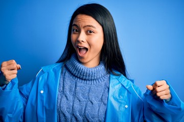 Wall Mural - Young beautiful chinese woman wearing rain coat standing over isolated blue background celebrating surprised and amazed for success with arms raised and open eyes. Winner concept.