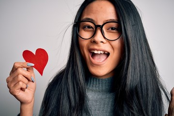 Wall Mural - Young beautiful chinese woman holding paper heart over isolated white background very happy and excited, winner expression celebrating victory screaming with big smile and raised hands