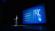 © Gorodenkoff - Live Event with Brand New Products Reveal: Speaker Presents Smartphone Device to Audience. Movie Theater Screen Shows Mock-up Touch Screen Mobile Phone with High-Tech Features and Top Highlights