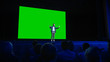 © Gorodenkoff - On Stage, Keynote Speaker Presents New Product to the Audience, Behind Him Movie Theater with Green Screen, Mock-up, Chroma Key. Business Live Event or Device Reveal