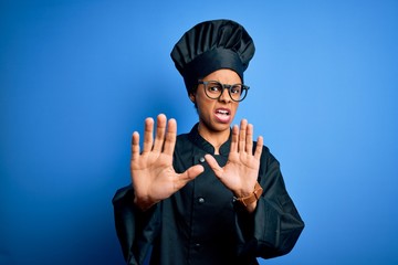 Wall Mural - Young african american chef woman wearing cooker uniform and hat over blue background afraid and terrified with fear expression stop gesture with hands, shouting in shock. Panic concept.