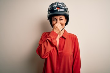 Poster - Middle age motorcyclist woman wearing motorcycle helmet over isolated white background smelling something stinky and disgusting, intolerable smell, holding breath with fingers on nose. Bad smell