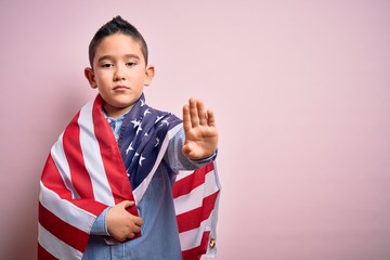 Poster - Young little patriotic boy kid covered on united states of america flag on independence day with open hand doing stop sign with serious and confident expression, defense gesture