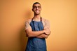 © Krakenimages.com - Young handsome african american shopkeeper man wearing apron over yellow background happy face smiling with crossed arms looking at the camera. Positive person.