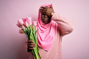 Poster - Young african american plus size woman wearing muslim hijab holding bouquet of pink tulips covering eyes with hand, looking serious and sad. Sightless, hiding and rejection concept