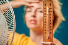A Portrait Of Woman In Front Of Fan Suffering From Heat Holding Thermometer. Close Up. Hot Weather Concept