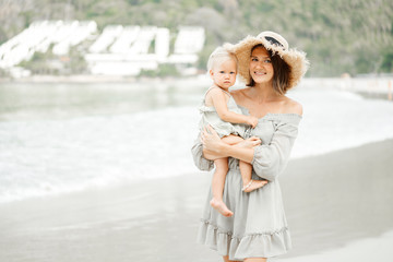 Young happy mother in a straw hat holds a blonde girl in the arms of her child and looks at the camera on the background of the beach and hotel. Tropical vacation.