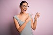 © Krakenimages.com - Young beautiful woman wearing casual striped dress and glasses over pink background smiling and looking at the camera pointing with two hands and fingers to the side.