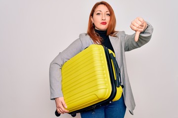 Canvas Print - Young beautiful redhead woman holding suitcase prepared to business travel with angry face, negative sign showing dislike with thumbs down, rejection concept