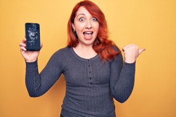 Poster - Young beautiful redhead woman holding broken smartphone showing cracked screen pointing thumb up to the side smiling happy with open mouth