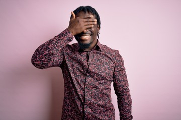 Poster - Young handsome african american man wearing casual shirt standing over pink background smiling and laughing with hand on face covering eyes for surprise. Blind concept.