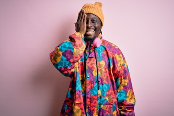 Canvas Print - Young handsome african american man wearing colorful coat and cap over pink background covering one eye with hand, confident smile on face and surprise emotion.