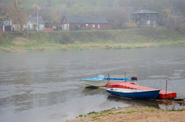 Sad autumn landscape. Fog and rain over the river. Rescue boats stand at the shore. Walk on the water in bad weather. Two boats moored at the river dock.