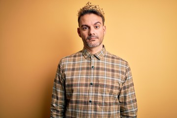 Young handsome man wearing golden crown of king over isolated yellow background with serious expression on face. Simple and natural looking at the camera.