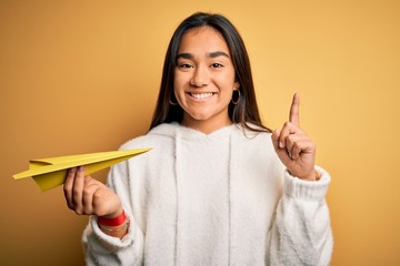 Canvas Print - Young beautiful asian woman holding paper airplane standing over isolated yellow background surprised with an idea or question pointing finger with happy face, number one