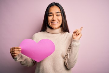 Canvas Print - Young beautiful asian woman holding pink heart standing over isolated background very happy pointing with hand and finger to the side