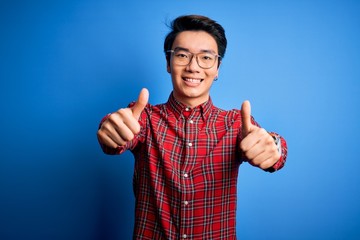 Young handsome chinese man wearing casual shirt and glasses over blue background approving doing positive gesture with hand, thumbs up smiling and happy for success. Winner gesture.