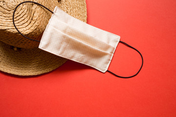 summer 2020 concept: straw hat and mask on plain red background. coronovirus covid holiday travel trip. risk of virus infection