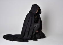 Fantasy Portrait Of A Woman With Red Hair Wearing Dark Leather Assassin Costume With Long Black Cloak.  Full Length Kneeling Pose, Isolated Against A Studio Background.