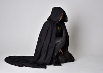 Fantasy portrait of a woman with red hair wearing dark leather assassin costume with long black cloak.  full length kneeling pose, isolated against a studio background.