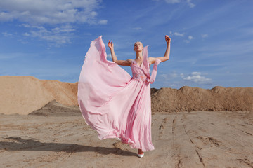 Horizontal view. Full length portrait of a young beautiful blonde high girl in fashionable flying pink maxi dress poses in sand quarry on sunny summer day.