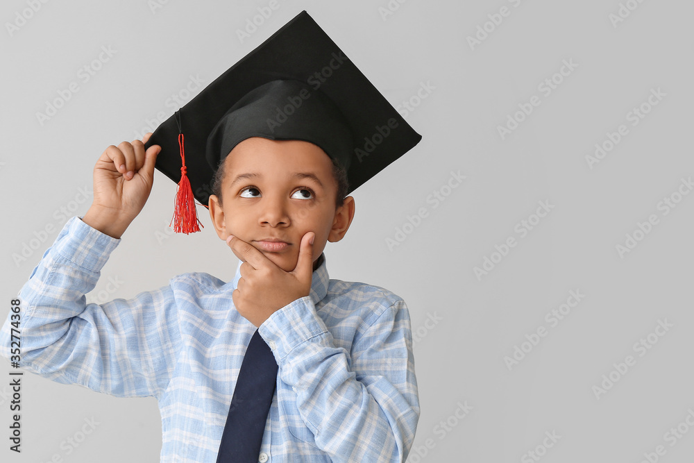 Thoughtful African-American boy in graduation hat on grey background
