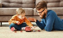 Side View Of Positive Young Bearded Man And Little Son Sitting On Floor Near Sofa And Playing The Toys Cars  While Enjoying Time Together At Home.
