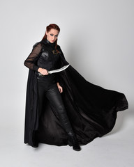 Canvas Print - fantasy portrait of a woman with red hair wearing dark leather assassin costume with long black cloak. Full length standing pose holding a sword isolated against a studio background.