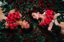 Two Brunettes Are Lying On The Flowers, Covered With Red Roses
