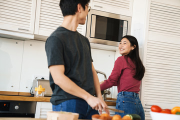 Wall Mural - Photo of joyful asian couple laughing and talking while cooking dinner