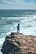 © Evgeniy - a young girl in a short white T-shirt with a backpack stands on the edge of a cliff and looks at the stormy sea in sunny windy clear weather. Journey and outdoor concept.