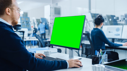 Over the Shoulder: Heavy Industry Engineer Working on Personal Computer, Display is Green Mock-up Screen or Chroma Key. Industrial Factory with High-Tech CNC Machinery, Professionals Working