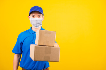 Wall Mural - Front of Asian young delivery worker man in blue t-shirt and cap uniform wearing face mask protective giving cardboard boxes under coronavirus or COVID-19, studio shot isolated yellow background