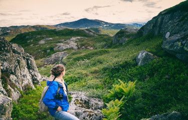 Wall Mural - Young woman traveler in a blue jacket with a backpack and a photo camera in the mountains, against the backdrop of a beautiful landscape