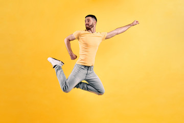 I am a superman! A young man dressed in a yellow T-shirt and light jeans jumps against a yellow wall, straightening one hand sideways like a superhero. Advertising concept.