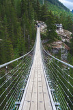 Fototapeta Mosty linowy / wiszący - Road on suspension foot bridge without people located in  BC Canada as landscape point view on another side of the rocks in the forest with green pine trees and nature view and adventure time