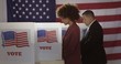 © vesperstock - Profile, medium shot, young mixed-race woman and Hispanic man in polling station, voting in a booth with US flag in background.
