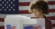 © vesperstock - MCU profile young, mixed race woman, alone casting vote at polling station. She looks down at ballot paper, out of frame. US flag in background.