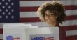 © vesperstock - MCU profile young, mixed race woman, alone casting vote in booth at polling station. She looks up towards viewer, smiling. US flag in background.