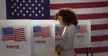Profile, Medium Shot, Young Hispanic Woman In Polling Station, Voting In A Booth With US Flag In Background.