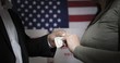 © vesperstock - CU detail of hands as anonymous man in shirt and jacket hands over cash to an unseen woman in front of voting booths and American flag.